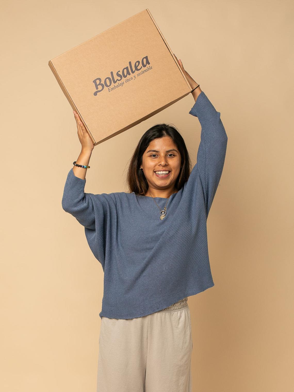 Caja automontable de cartón kraft con el logo de Bolsalea sostenida por una mano sobre una mesa con fondo verde. Embalaje ético y sostenible fabricado en España con empleo inclusivo y materiales reciclables.