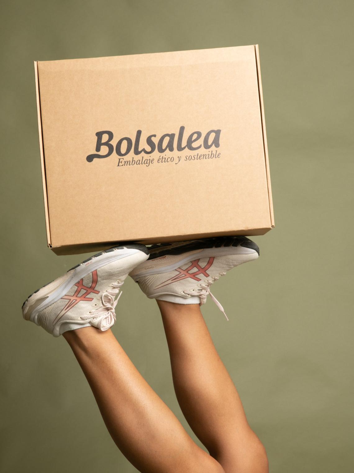Piernas levantadas sosteniendo una caja automontable de cartón kraft con el logo de Bolsalea. Embalaje ético y sostenible fabricado en España, reciclable y fruto del empleo inclusivo. Ideal para envíos de moda, accesorios o ecommerce responsable.