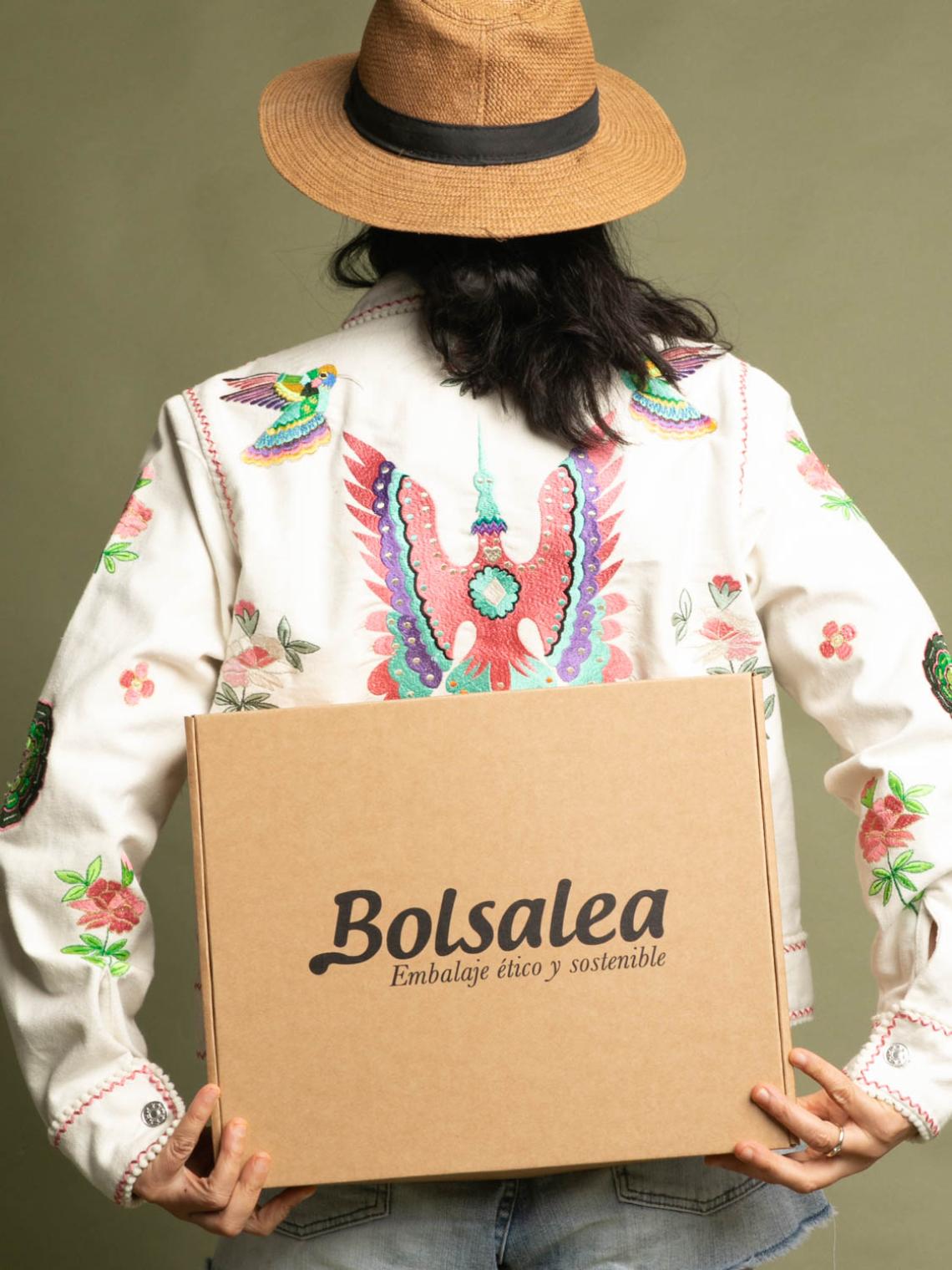 Persona con sombrero y chaqueta bordada sosteniendo una caja automontable de cartón kraft con el logo de Bolsalea. Embalaje ético y sostenible fabricado en España con impacto social. Si deseas tu caja con separadores interiores, contáctanos.