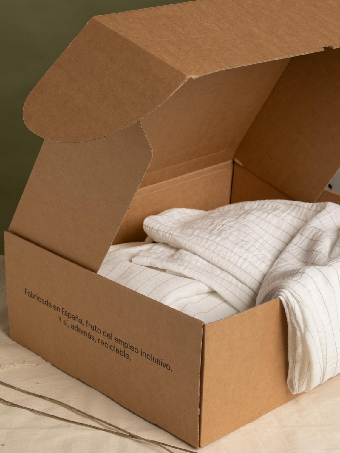 Caja automontable de cartón kraft abierta, con una prenda textil blanca en su interior. En el lateral se lee “Fabricada en España, fruto del empleo inclusivo. Y sí, además, reciclable.” Embalaje sostenible ideal para envíos de moda o textil.