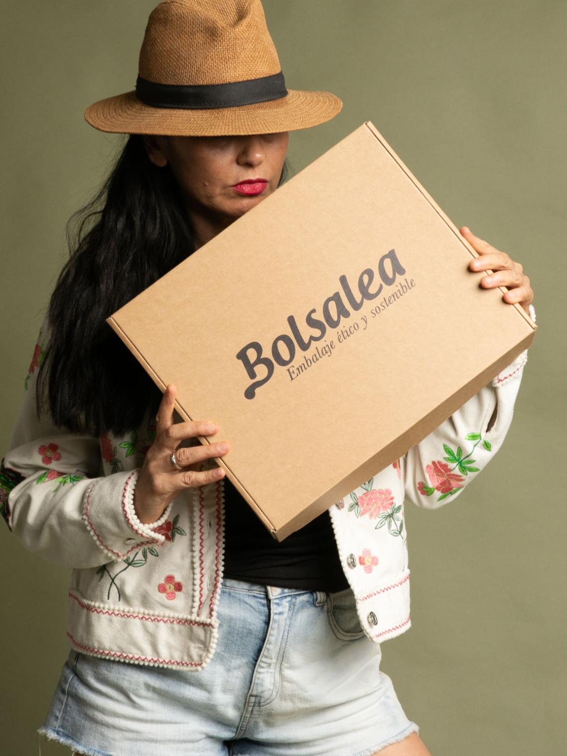 Persona con sombrero y chaqueta bordada sosteniendo una caja automontable de cartón kraft con el logo de Bolsalea. Embalaje ético y sostenible fabricado en España con impacto social. Si deseas tu caja con separadores interiores, contáctanos.