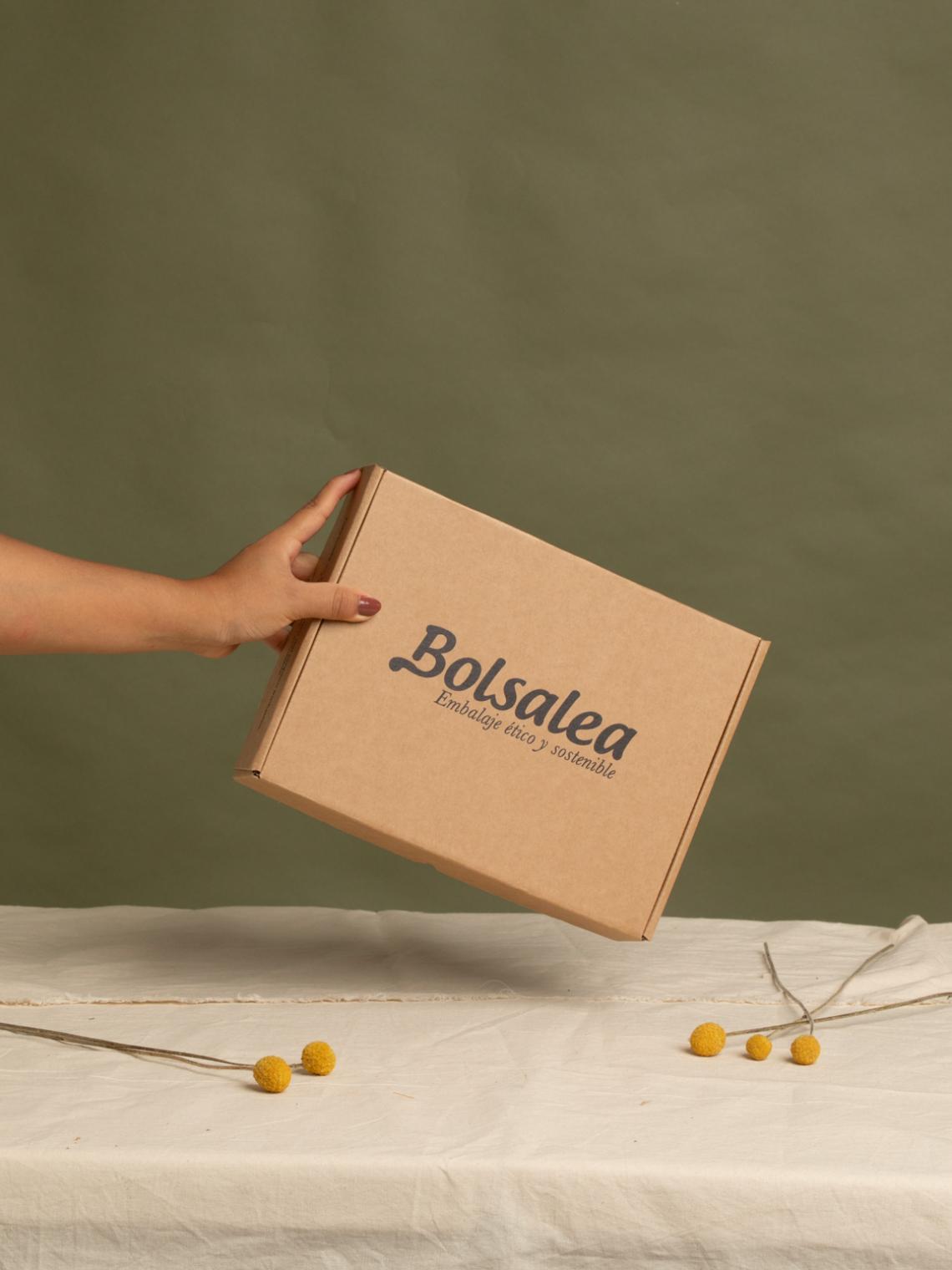 Caja automontable de cartón kraft con logo de Bolsalea | Packaging sostenible Mano sosteniendo una caja automontable de cartón kraft con el logo de Bolsalea y el lema “Embalaje ético y sostenible”. Fabricada en España con impacto social. Si deseas tu caja con separadores interiores, contáctanos.