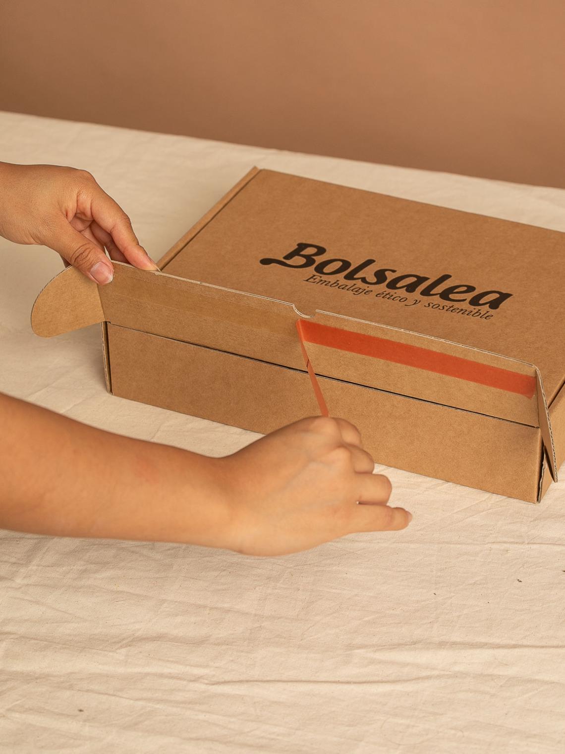 Manos cerrando una caja automontable de cartón kraft con el logo de Bolsalea y cinta adhesiva naranja. Embalaje ético y sostenible fabricado en España con impacto social. Si deseas tu caja con separadores interiores, contáctanos.