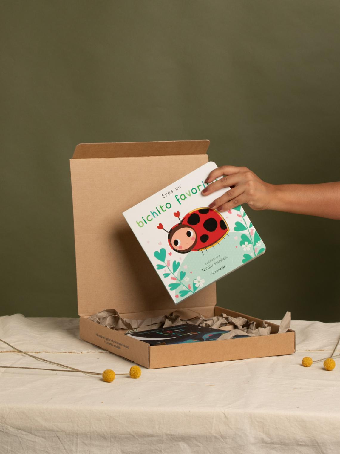 Caja automontable de cartón kraft abierta con una mano colocando un libro infantil en su interior. Fabricada en España con empleo inclusivo y materiales reciclables, ideal para packaging de libros o regalos sostenibles.