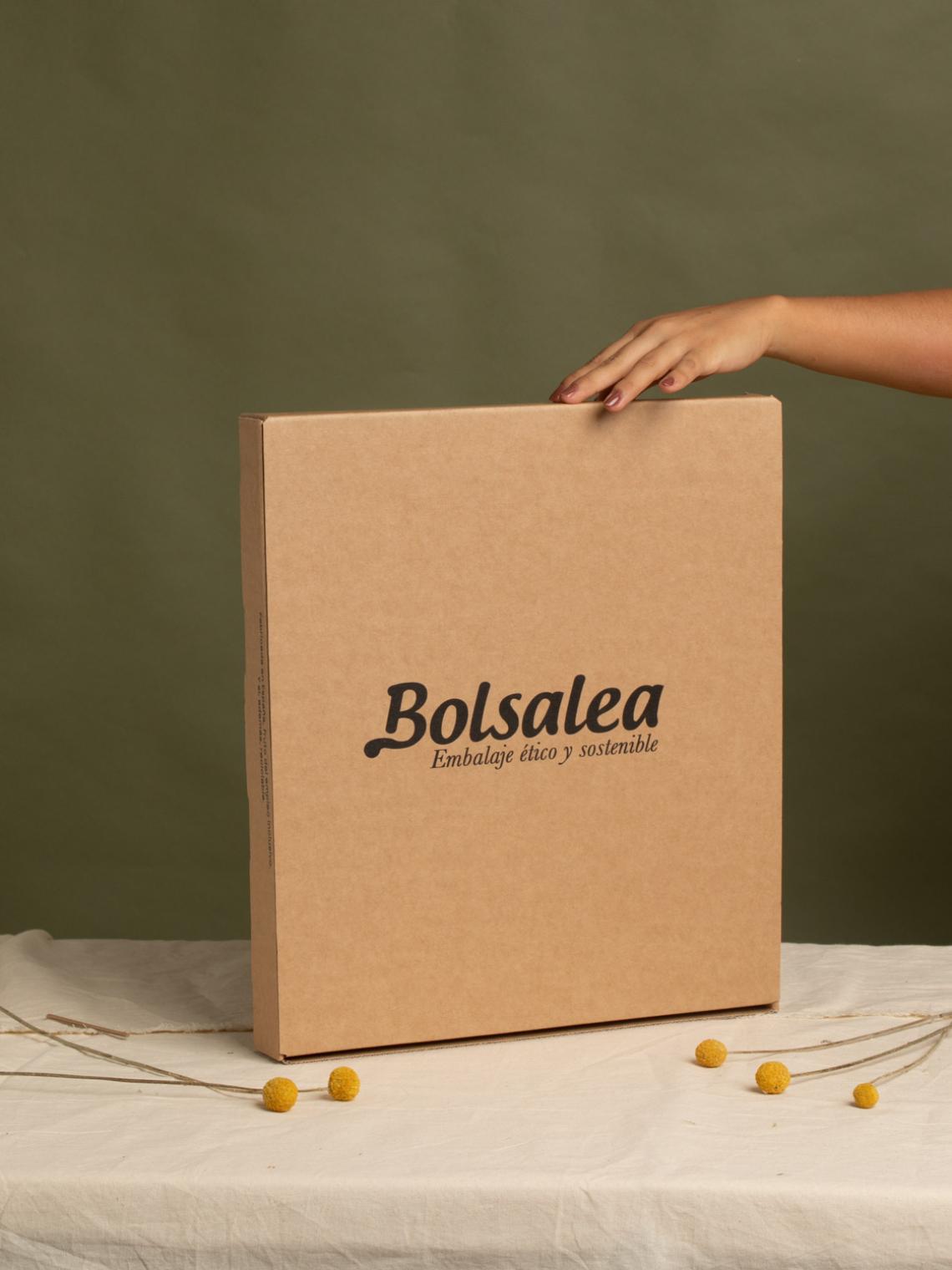 Caja automontable de cartón kraft para envíos | Embalaje sostenible Bolsalea Caja automontable de cartón kraft con logo de Bolsalea sobre fondo verde y decoración floral amarilla. Embalaje ético, reciclable y fabricado en España con empleo inclusivo.